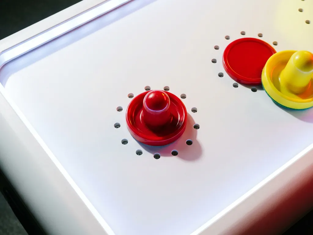 Rode air hockey puck op wit tafelvlak met tweede puck en gele slagmallet, luchtvents zichtbaar, bovenaanzicht.