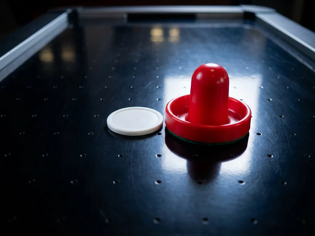 Rode luchthockeyslagschijf en witte puck op glanzend zwart luchthockeytafel oppervlak met zichtbare luchtgaatjes.