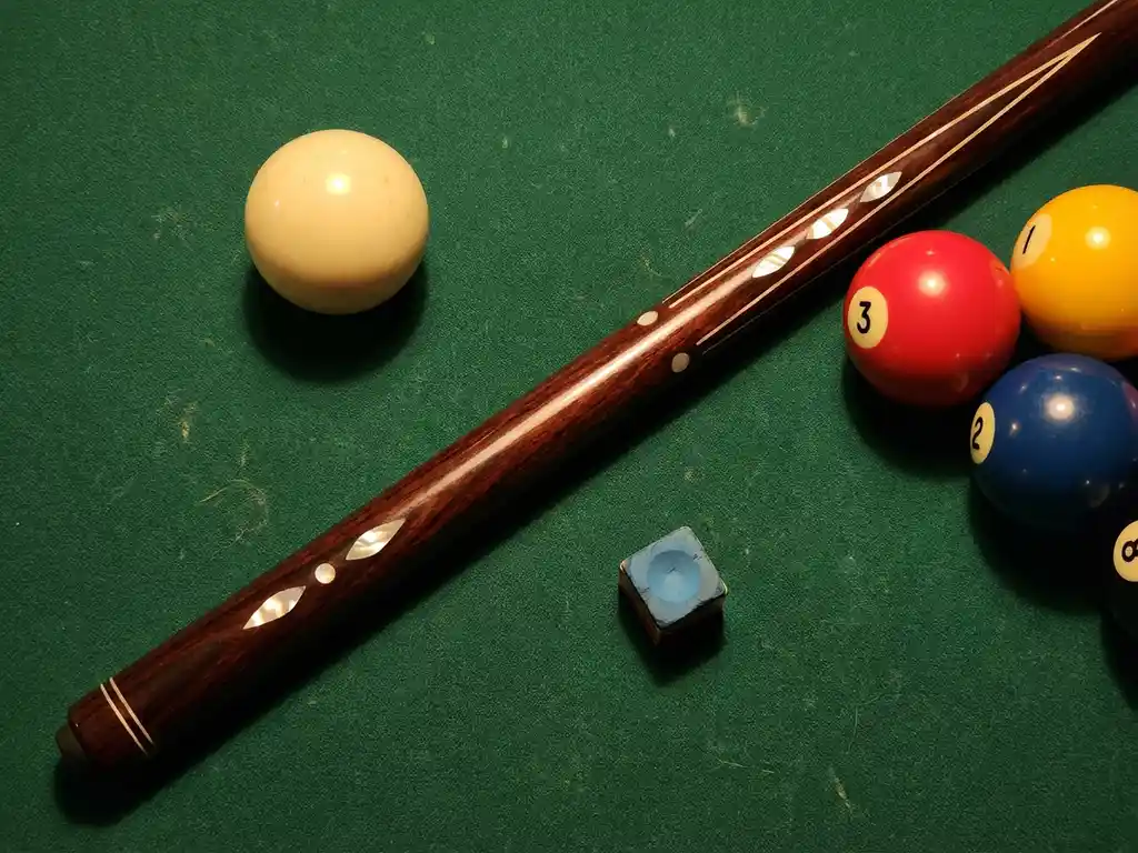 Premium biljartqueue diagonaal op groen laken met ivoren speelbal, gekleurde ballen en krijtblok.