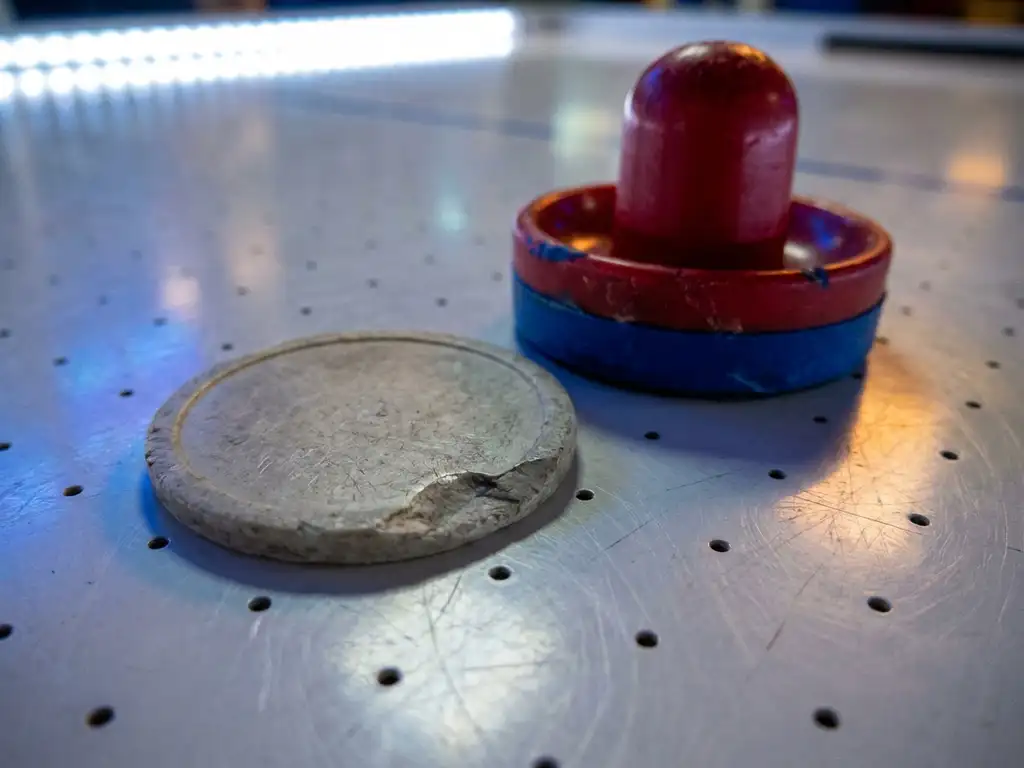 Versleten luchthockeypuck en slagmallet op wit speeloppervlak van een luchthockeytafel, met zichtbare krassen en afgebrokkelde rand.