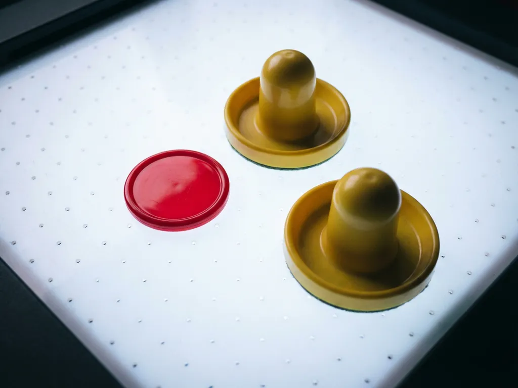 Rode luchthockeypuck en twee gele slagpaddles op wit speelvlak van een luchthockeytafel, gefotografeerd van bovenaf.