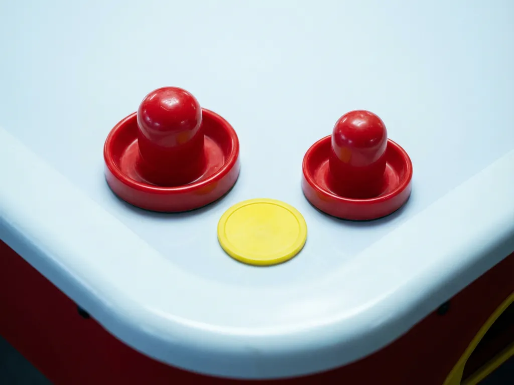 Twee rode luchthockeyslagjes van verschillend formaat naast elkaar op een wit tafeloppervlak, met een gele puck ertussenin.