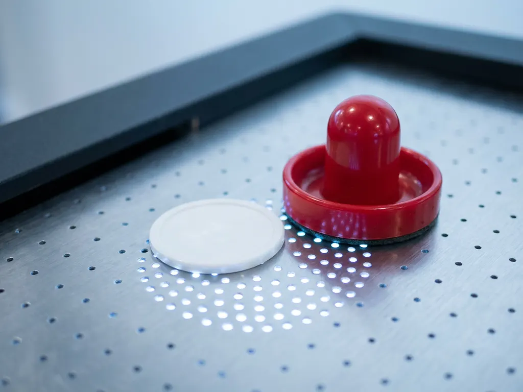 Rode air hockey-striker en witte puck op geperforeerd speeloppervlak met zichtbare luchtgaten in macro-close-up.