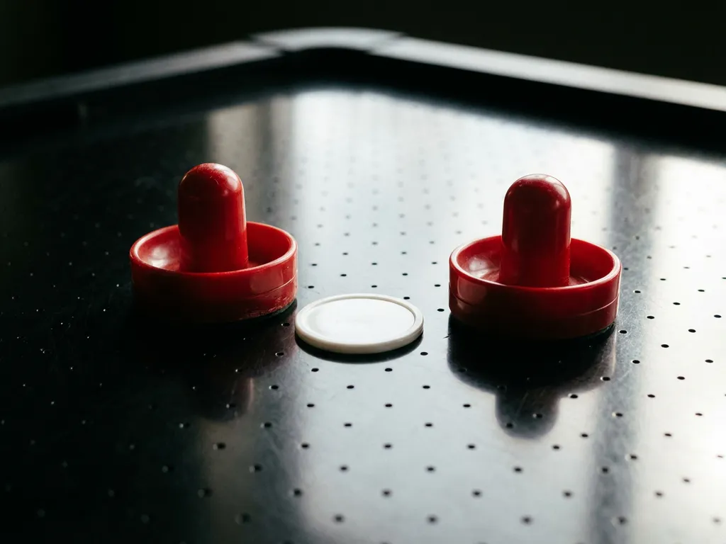 Twee rode air hockey strikers en een witte puck op een glanzend zwart speelveld met zichtbare luchtgaatjes.