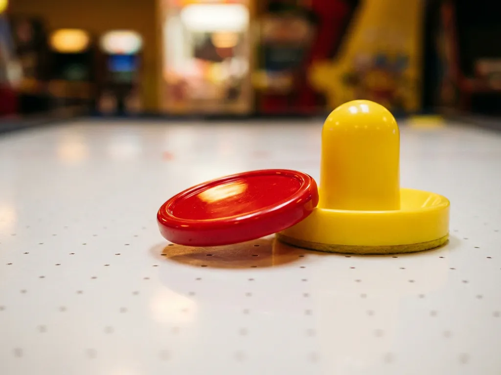 Rode luchthockeypuck zwevend boven wit speeloppervlak met zichtbare luchtgaatjes, gele slagmallet ernaast.